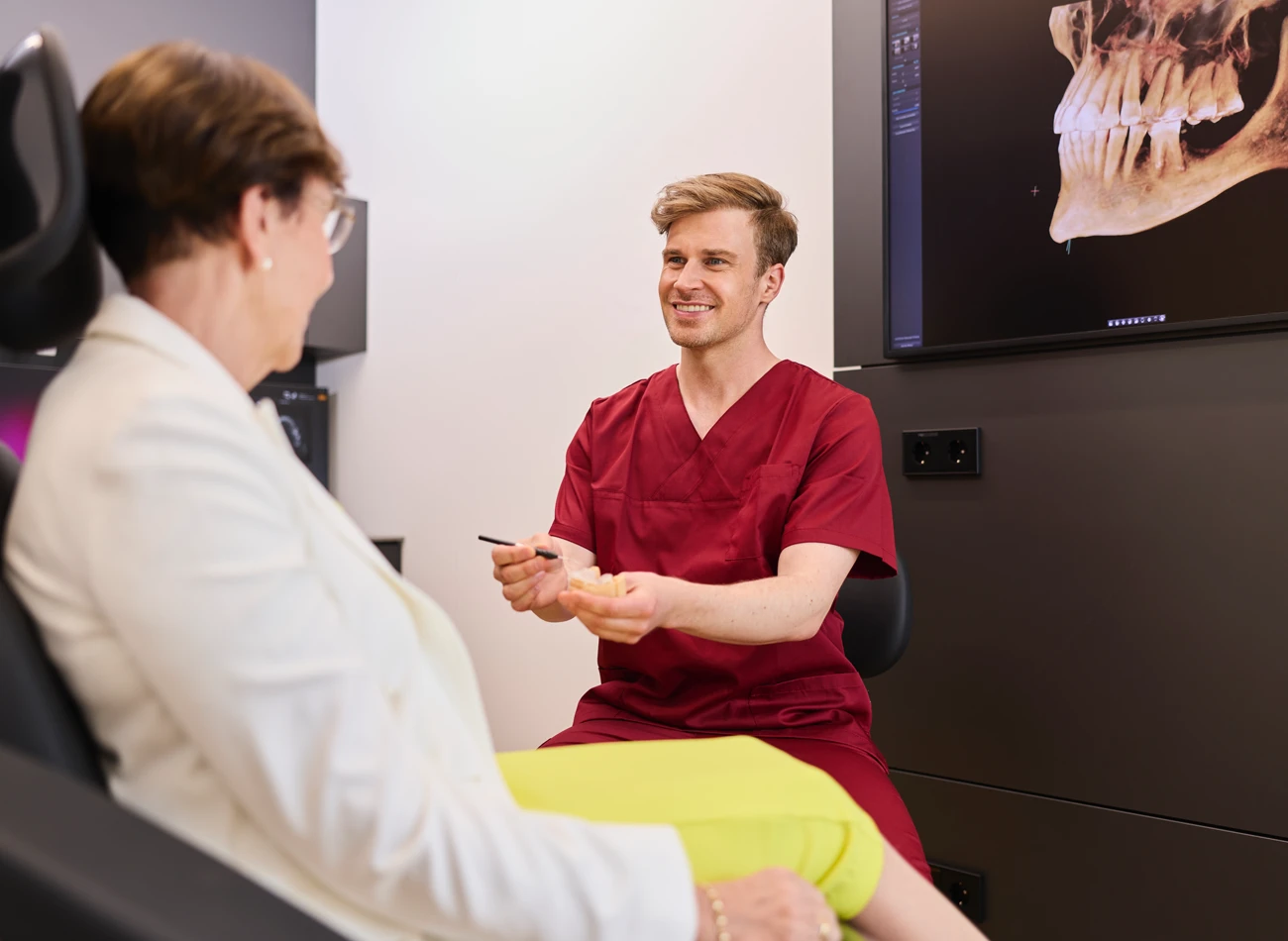 Fachzahnarzt Philipp Deuringer im Beratungsgespräch mit Patientin zum Thema All-on-4. Patientin sitzend im Behandlungsstuhl, Herr Deuringer zeigt ihr ein Zahnmodell.